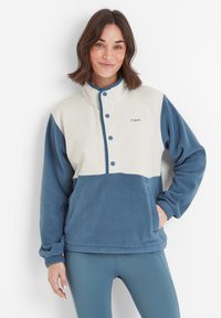 Smiling woman wearing a blue and white fleece jacket with snap buttons, standing with one hand in pocket against a white background.