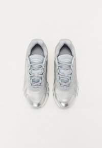 Sapatilhas cinzentas com um revestimento prateado brilhante, lingueta em malha, atacadores cinzentos e um design de sola ondulante distintivo. Apresentam a marca da Nike na lingueta.