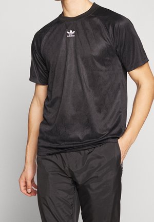 Męski tors w czarnej koszulce Adidas z krótkim rękawem z subtelnym geometrycznym wzorem i czarnych spodniach, jedna ręka w kieszeni.