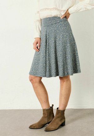 Woman wearing a knee-length blue floral skirt, white long-sleeve blouse, and brown suede ankle boots, standing with one hand on hip.