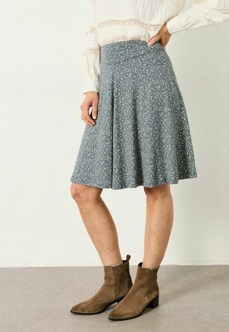 Naine, kes kannab põlveni ulatuvat sinist lillelist seelikut, valget pikkade varrukatega pluusi ja pruune seemisnahast põlviksaapaid, seisab ühe käega puusal.