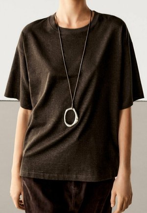 Personne portant une chemise ample à manches courtes marron foncé et un long collier avec un grand pendentif ovale en argent.