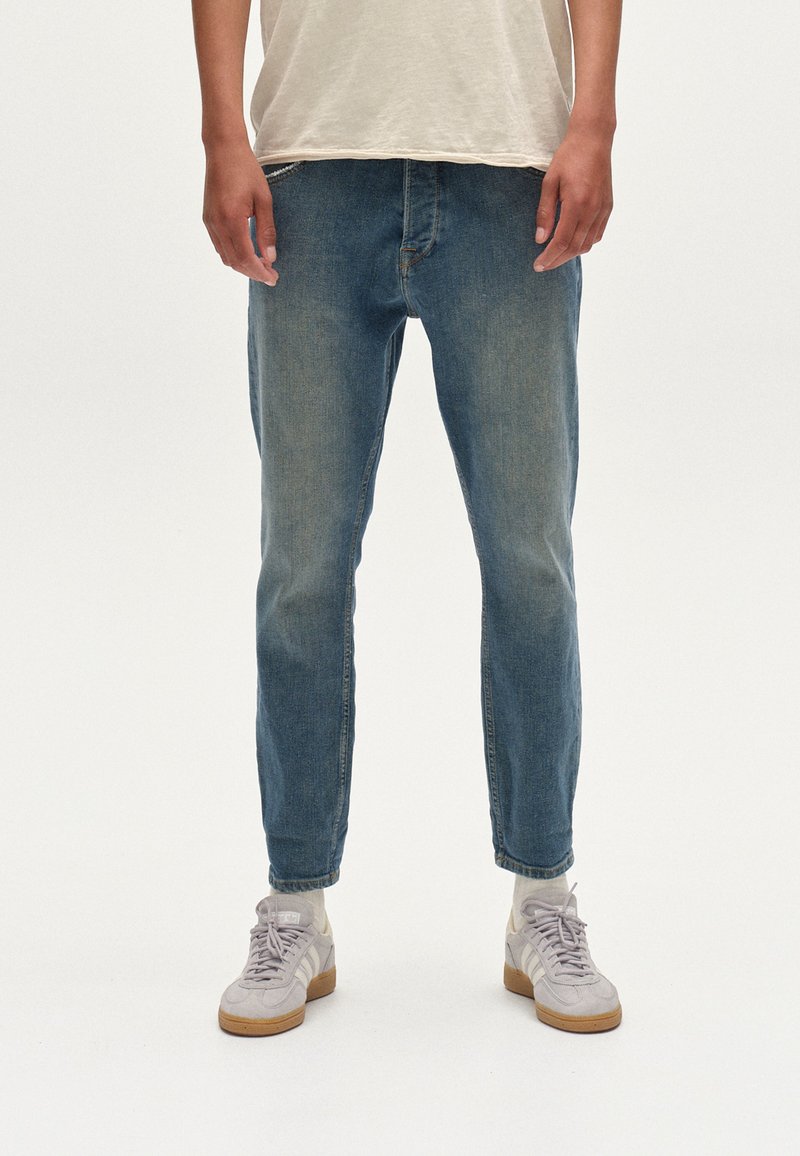 Világoskék denim farmer slim fit szabással, enyhén szűkült lábszárral és felhajtott szegéllyel. Bézs sportcipővel és egy egyszerű világos színű pólóval viselve.