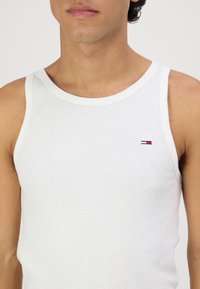 Top sin mangas de algodón blanco con escote redondo, que presenta un pequeño logo multicolor en la parte izquierda del pecho. Textura suave y diseño ajustado.