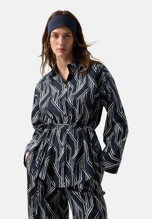 Frau trägt passende schwarze und weiße Kleidung mit geometrischem Muster, bestehend aus Hemd und Hose, dazu ein breites schwarzes Stirnband, steht mit den Händen in den Taschen.