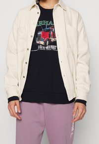 Chaqueta de camisa de corduroy color beige claro con botones a presión, llevada sobre una camiseta negra con un diseño de camión rojo. También incluye pantalones de chándal lavanda.
