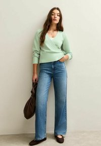 Suéter envolvente verde menta, jeans de pierna ancha azul claro, zapatos planos marrones y bolso de mano marrón oscuro sobre un fondo neutro.