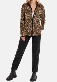 Kobieta w bluze polarowej z nadrukiem lamparta, czarnych dżinsach i czarnych botkach za kostkę, stoi z jedną ręką w kieszeni kurtki.