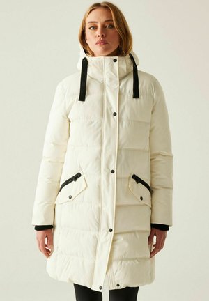 Kobieta ubrana w długi biały, ocieplany zimowy płaszcz z kapturem i czarnymi kieszeniami na zamek, stojąca na jednolitym jasnym tle.