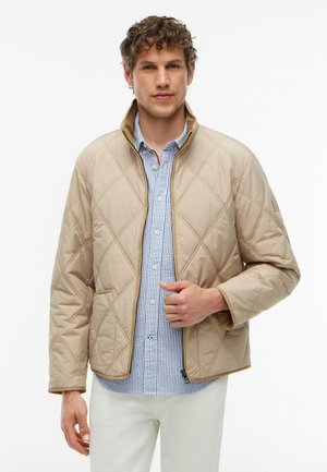 Ung mand iført en beige quiltet jakke over en blå- og hvidstribet skjorte og hvide bukser, stående mod en hvid baggrund.