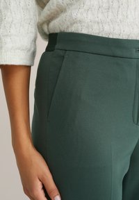 Tuva cilvēka tuvplāns, kurš valkā tumši zaļas bikses ar sānu kabatām un krēmkrāsas adītu džemperi, roka atslābusies tuvu gurnam.