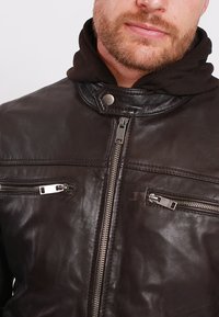 Černá kožená bunda s vysokým límcem, předním zipem a dvěma zipovými kapsami na prsou. Měkká textura a elegantní design.