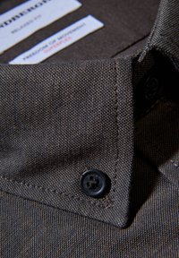 Brun texturerad tygskjortkrage med svarta knappar och skötselrådsetiketter. Närbild som framhäver sömnadsdetaljer och materialets väv.