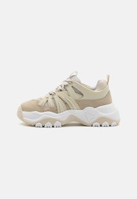 Beige og krem fottøy til sport med mesh-paneler, en teksturert semsket overdel, og en kraftig hvit såle med et karakteristisk mønster.