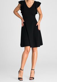Vestido negro con escote en V y mangas con volantes. Falda acampanada con una textura suave. Combinado con tacones negros de tiras.