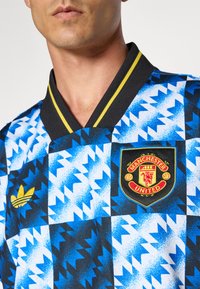 Adidas Manchester United-Trikot mit einem blau-schwarzen geometrischen Muster, das mit gelben Akzenten und einem gestickten Logo auf der Brust versehen ist.