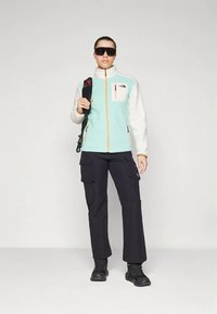 Vildist jakk mintrohelises ja valges oranži luku ning mustade lahtiste pükstega matkasaabastega. Käepärast seljakott.