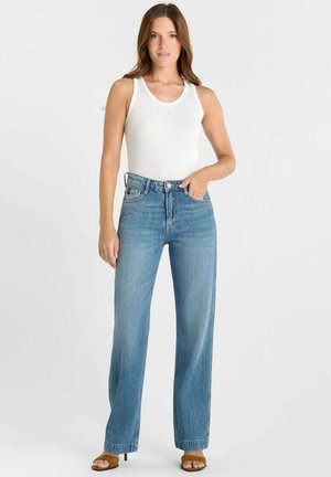 Wit tanktop met een ronde halslijn, gecombineerd met lichtblauwe, hooggetailleerde wijde jeans. Bruine sandalen met hak maken de outfit compleet.