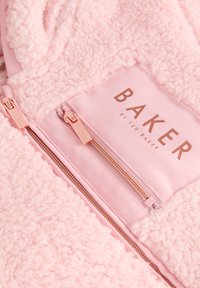 Primo piano di una giacca in tessuto rosa strutturato con due cerniere in oro rosa e un'etichetta "BAKER by Ted Baker".