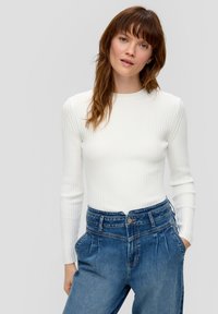 Un haut blanc à longues manches côtelé associé à un jean bleu taille haute, avec des plis et des poches, mettant en avant un design ajusté et texturé.