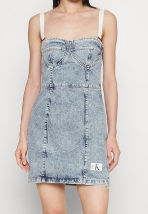 Femme portant une mini-robe en denim bleu délavé avec des bretelles blanches réglables et une petite étiquette logo Calvin Klein près de l'ourlet.