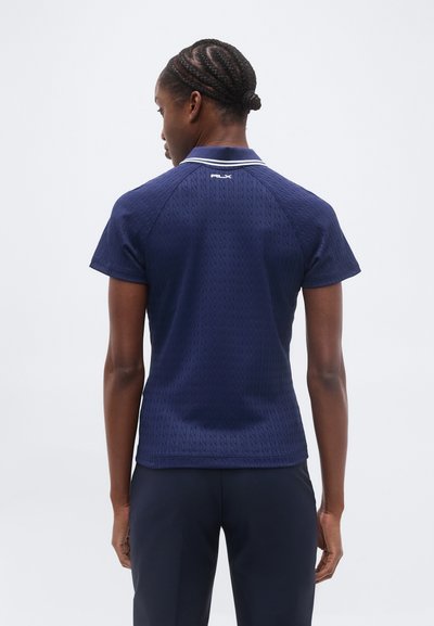 Γυναίκα με πλεγμένα μαλλιά, φορώντας μπλε ναυτικό πόλο με κοντά μανίκια και σκούρο παντελόνι, γυρισμένη προς τα πίσω, μπροστά σε απλό φωτεινό φόντο.