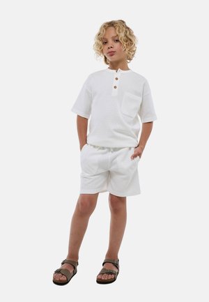 Chemise henley blanche à manches courtes avec trois boutons en bois et une poche, associée à un short blanc assorti, portée avec des sandales brunes.