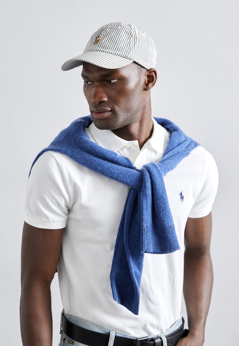 Camisa polo branca com um pequeno logótipo, suéter azul atado ao pescoço e um boné riscado. As texturas notáveis incluem lã e algodão.