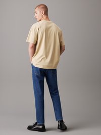 Beige bomullströja med avslappnad passform och rundad halsringning, i kombination med blå denimjeans och svarta snörskor. Enkel, avslappnad design.