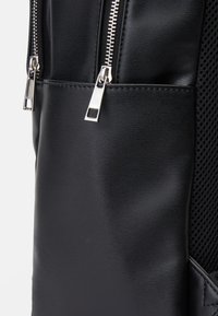 Sac à dos noir en matériau synthétique lisse et texturé, avec des zips argentés et un accent en maille sur le côté. Design compact et minimaliste.