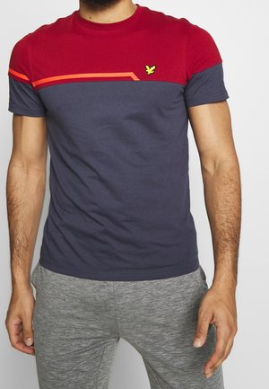 Homme portant un t-shirt à manches courtes rouge et bleu marine avec un design en blocs de couleurs, arborant un petit logo d'oiseau jaune sur la poitrine.