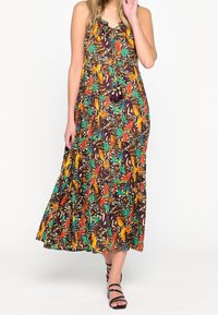 Žena v bezrukávových maxi šatech s barevným tropickým potiskem a třásněmi, doplněná černými sandály na podpatku se svázanými pásky, na bílém pozadí.
