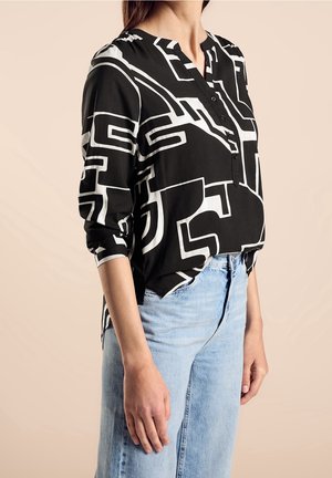 Frau trägt eine schwarz-weiß geometrisch gemusterte Langarmbluse mit Schlitz am Ausschnitt und hellblaue, hoch taillierte Jeans.