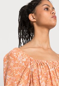 Top floral laranja com decote quadrado, detalhes franzidos e um tecido leve. Apresenta um padrão de flores delicadas.