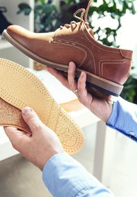 Brown leather shoe with perforated design and contrasting stitching, held above a removable cork insole with perforations, on a light surface.
