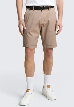 Man draagt beige shorts met een zwarte riem, witte sokken en witte sneakers, staand tegen een effen achtergrond.