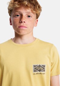 Žluté bavlněné tričko s černobílým grafickým logem na levé straně hrudi. Jednoduchý kulatý výstřih a krátké rukávy.