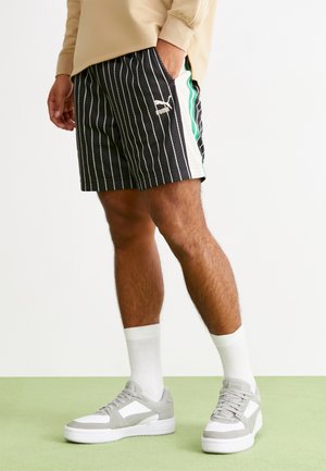 Zwarte short met krijtstreep en gaasstructuur, witte zijpanelen en een groene accentstreep; gecombineerd met grijze en witte sneakers en witte sokken.