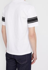 Valkoinen poolopaita mustilla raidoilla hihoissa, valmistettu puuvillasta, klassisella kauluksella ja lyhyillä hihoilla, yhdistetty tummiin farkkuihin.