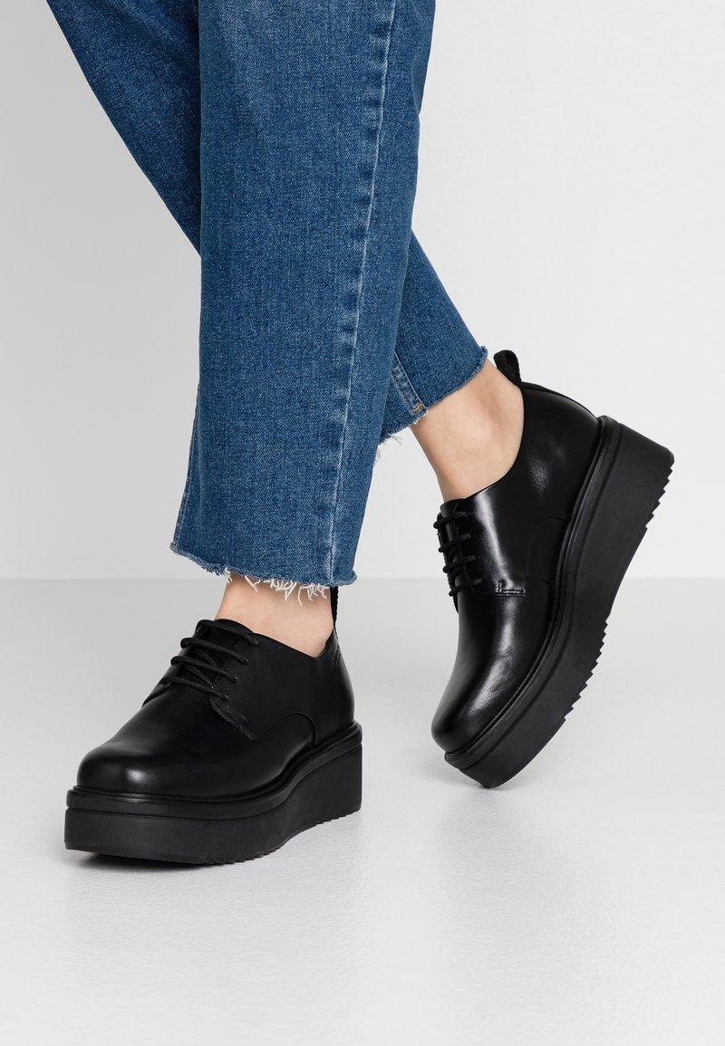 Personne portant un jean bleu et des chaussures à plateforme noires à lacets, debout sur un sol gris clair devant un fond uni.