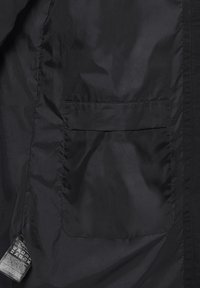 Schwarzes Stoffjacken-Innenfutter mit eingenähter rechteckiger Tasche und Pflegeetikett, das in die linke Seitennaht eingearbeitet ist.