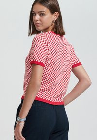 Suéter de manga corta a cuadros rojos y blancos con un dobladillo y cuello de canalé rojo. Textura suave, forma ajustada, combinado con pantalones oscuros.