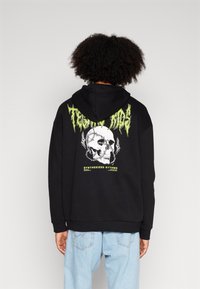 Sudadera negra con un gráfico de calavera y texto verde en la parte posterior. Cuenta con capucha con cordón y corte relajado. Material de algodón.