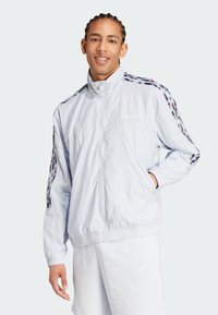 Hellblaues Windbreaker-Jacke aus leichtem Stoff, mit elastischen Bündchen, Seitentaschen und einem bedruckten Streifendesign auf den Schultern.