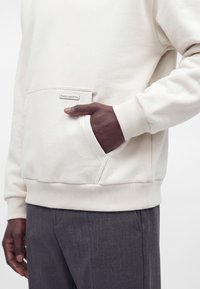 Personne à la peau foncée portant un sweat crème avec une poche à l'avant, main reposant dans la poche, associée à un pantalon gris.
