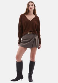 Brauner V-Ausschnitt-Pullover, grauer Wickelrock, schwarze kniehohe Stiefel. Weiche Textur, lockerer Schnitt, goldener Gürtelakzent, minimalistisches Design.