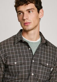 Bruine geruite shirt met een kraagontwerp, voorzien van twee voorzakken, knoopdetails en een gestructureerde basislaag in lichtgroen.