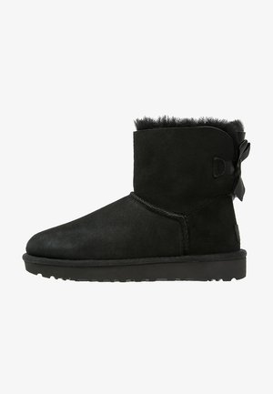Black suede ankle boot with soft fur lining and a flat rubber sole. Features a ribbon accent at the back and a rounded toe design.