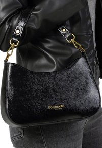 Sac à main en simili cuir noir avec une surface texturée, une forme incurvée, des accents en métal doré et un logo à l'avant. La bandoulière est réglable.