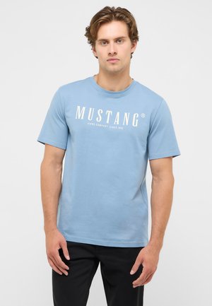Junger Mann mit welligem hellbraunem Haar, der ein hellblaues T-Shirt mit Mustang-Logo und schwarze Hose trägt, steht vor einem einfarbigen Hintergrund.